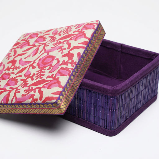 A square decorative box with a patterned pink and red floral lid leaning open next to a purple woven basket, set on artificial green grass against a dark blue backdrop.