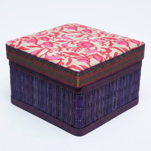 A square decorative box with a patterned red and white lid and a dark purple woven base, displayed on artificial green grass against a dark blue backdrop.